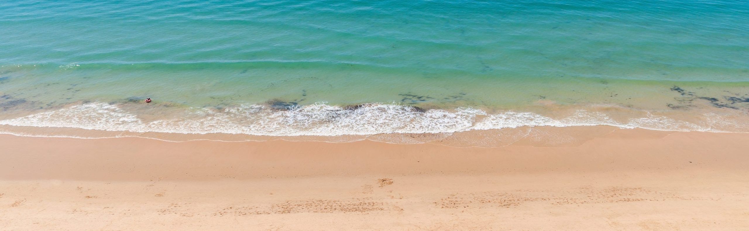 Imagen desde arriba de una playa con un barco a lo lejos, olas en la orilla, gente al sol y sombrillas de colores.